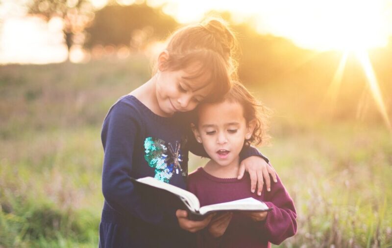 girl's left hand wraps around toddler while reading a book during golden hour